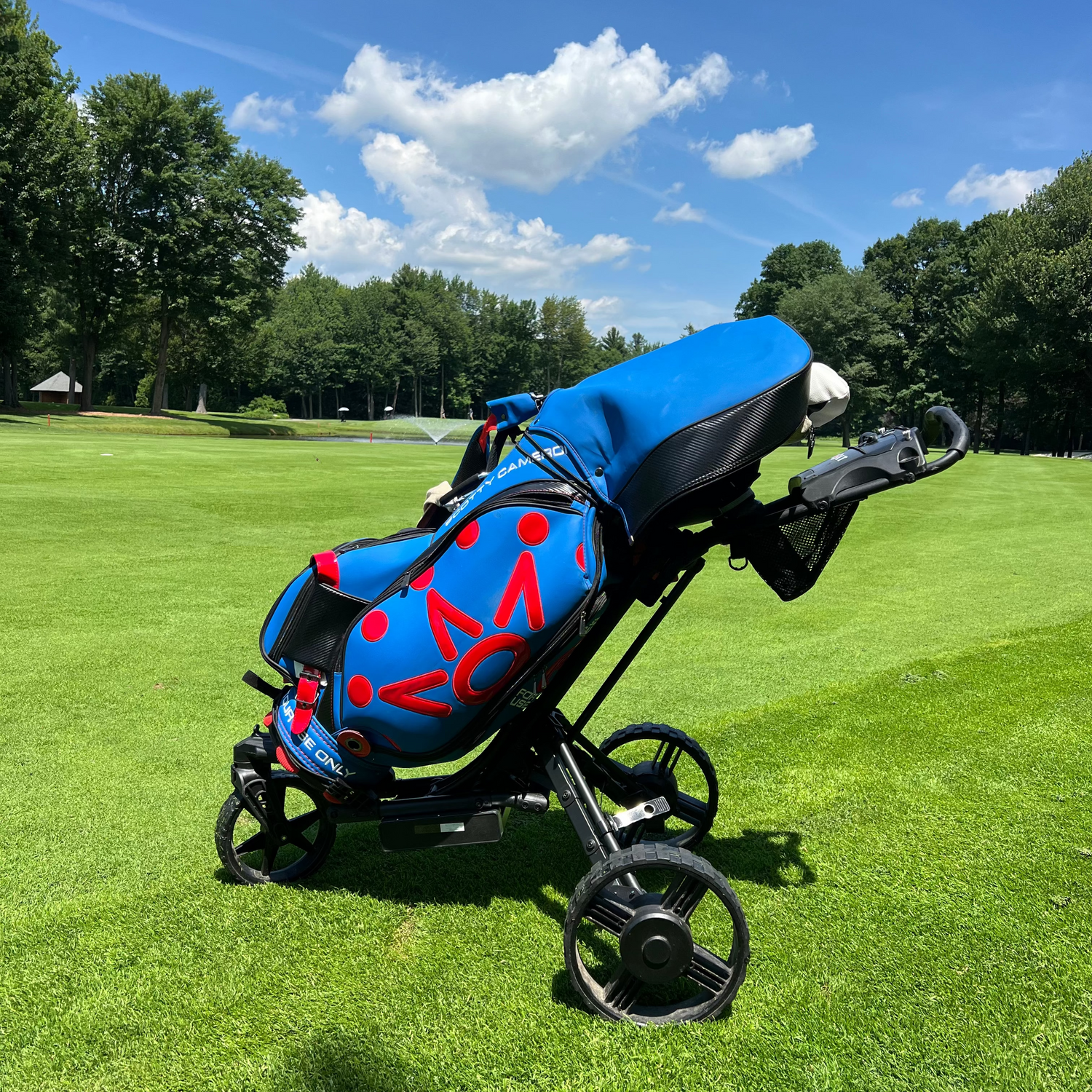 A blue and red golf stroller with a black frame is parked on a green grassy golf course under a blue sky with white clouds.
